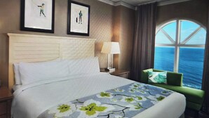 A hotel room with a large bed, a nightstand, a lamp, two framed pictures on the wall, a window with a view of the sea, and a green armchair. - Marriott OceanWatch 2-bedroom villa in Myrtle Beach, SC (Myrtle Beach)
