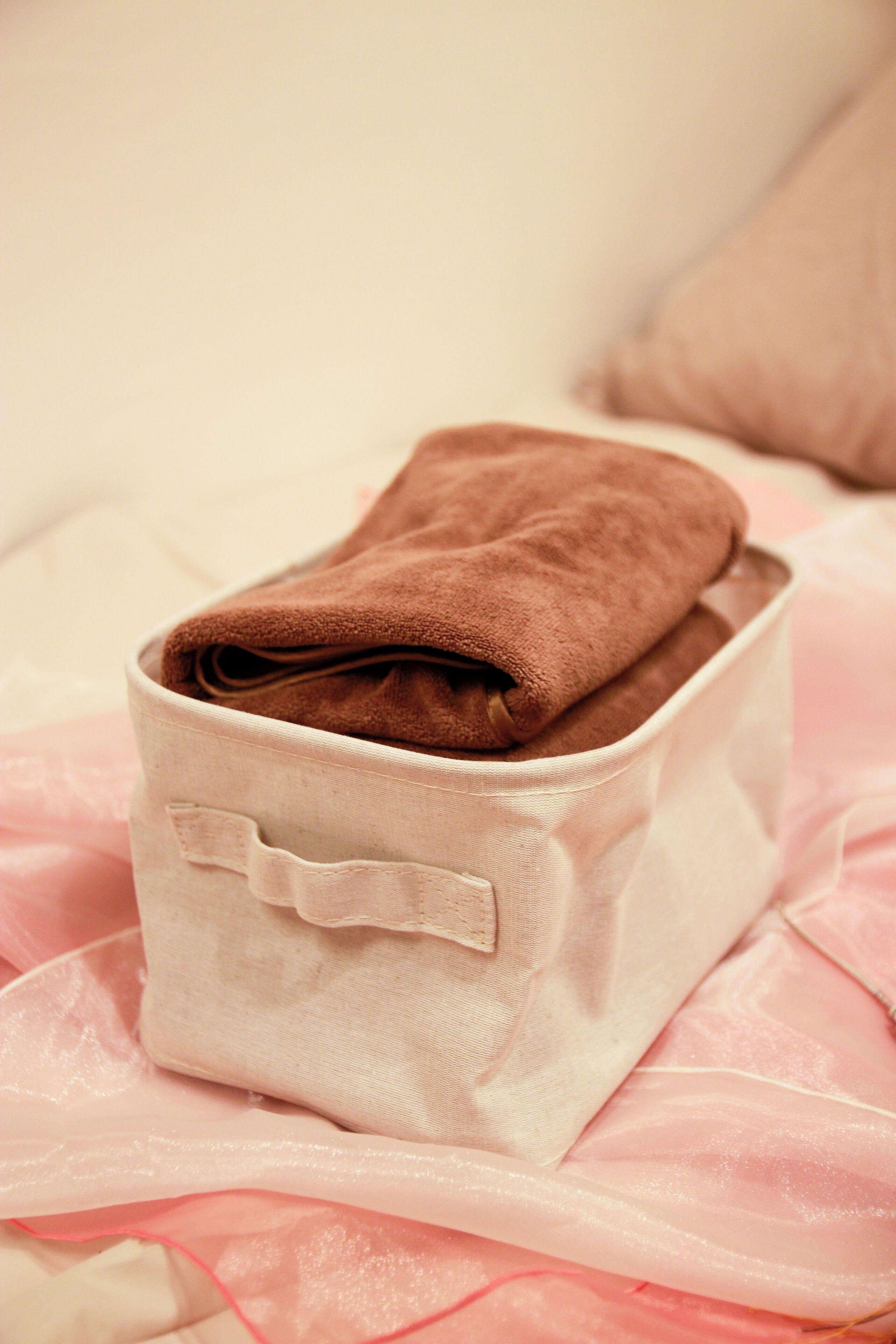 Una caja de tela color beige con un lazo decorativo en la parte frontal, colocada sobre una cama con colcha rosa.