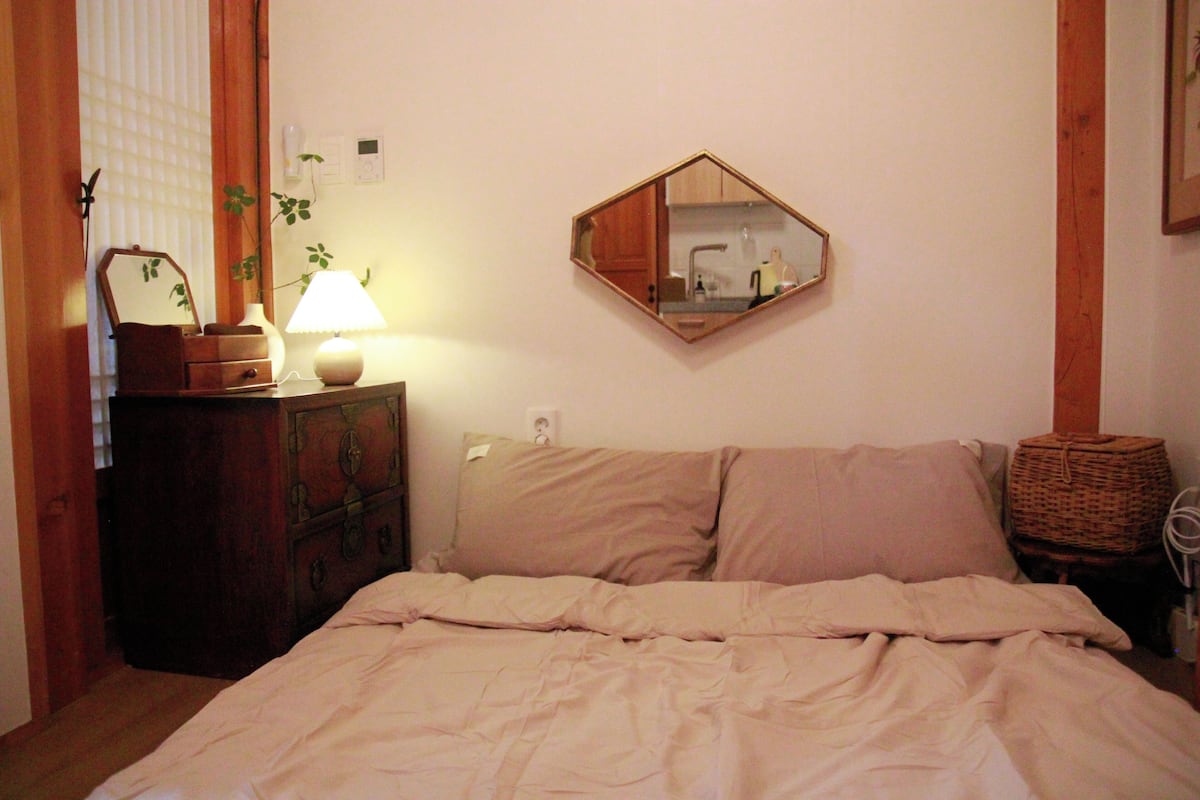 A bedroom with a bed, a dresser, a lamp, a mirror, and a plant.