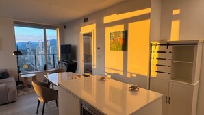 A modern kitchen with a large island, a dining area with a table and chairs, a sofa, and a view of a cityscape through large windows.