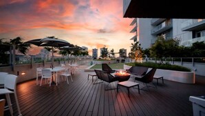 A rooftop terrace with outdoor seating, a fire pit, and a view of the city skyline during sunset.