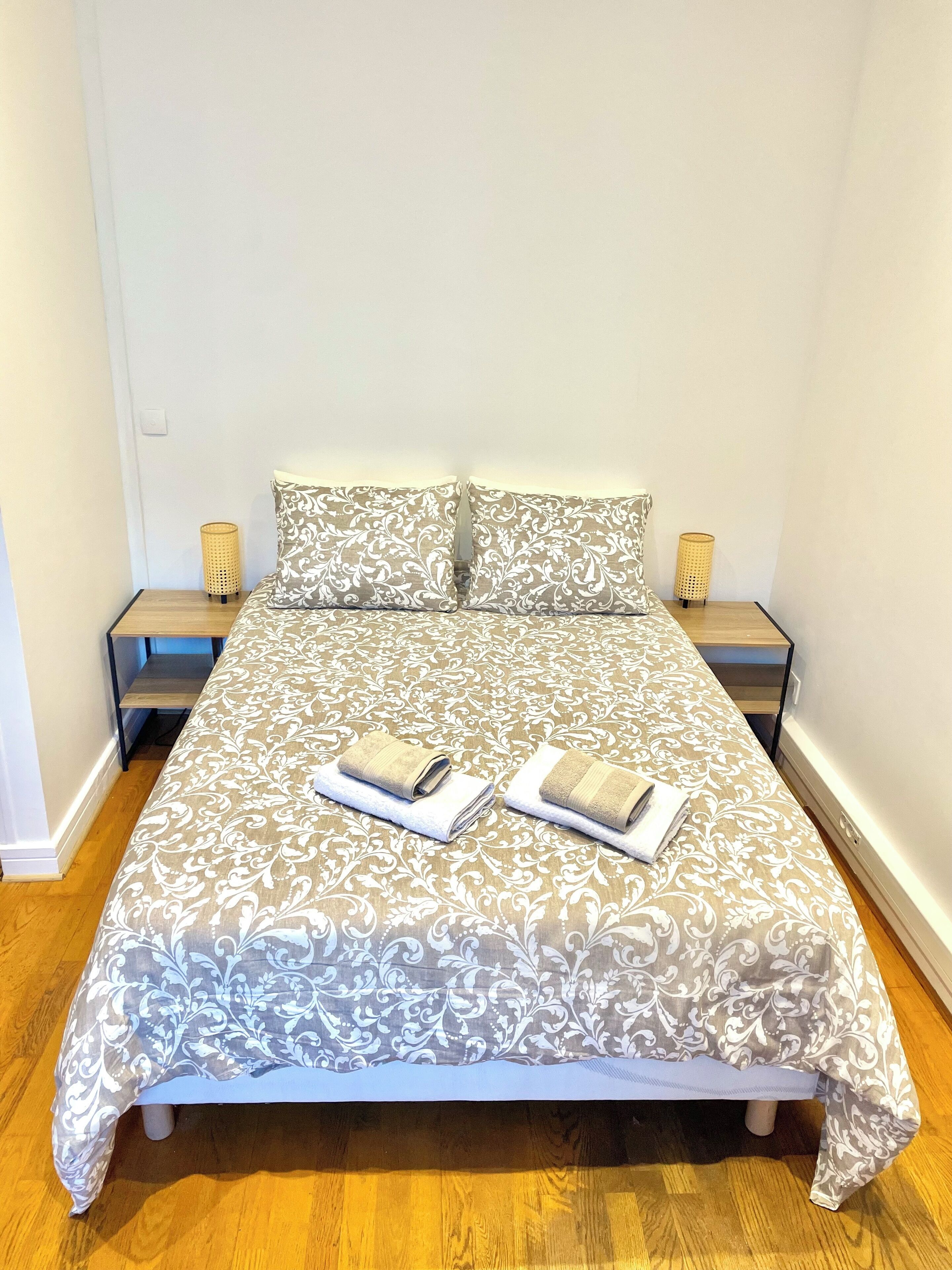 Un lit bien fait, recouvert d’une couvre-lit à motifs, avec deux oreillers et deux serviettes pliées posées dessus. De chaque côté du lit, on trouve une table de chevet sur laquelle est posée une lampe.