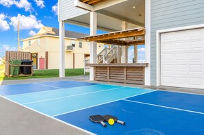 Ein Wohnbereich im Freien mit einem blauen Tennisplatz, einer Holzterrasse und einer Garage.