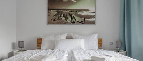 Ein Hotelzimmer mit einem Bett, weißen Leinentüchern, zwei Nachttischen mit Lampen und einem gerahmten Bild mit Strandmotiv an der Wand.