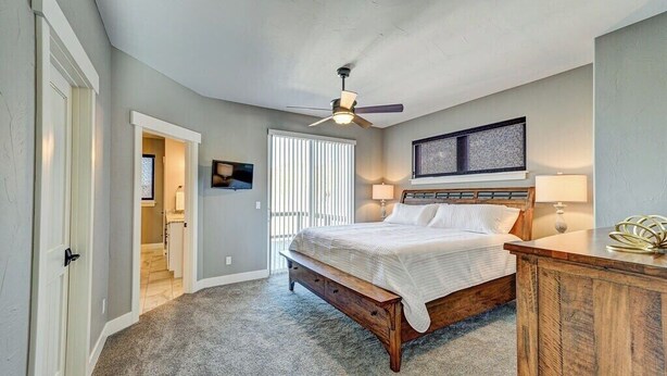 Quarto com cama, ventilador de teto, janela com persianas e porta que dá para outro cômodo.