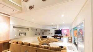 Ein modernes Wohnzimmer mit einer Couch, Sesseln und einem Fernseher. Regale mit dekorativen Gegenständen und eine Glastür, die nach draußen führt.
