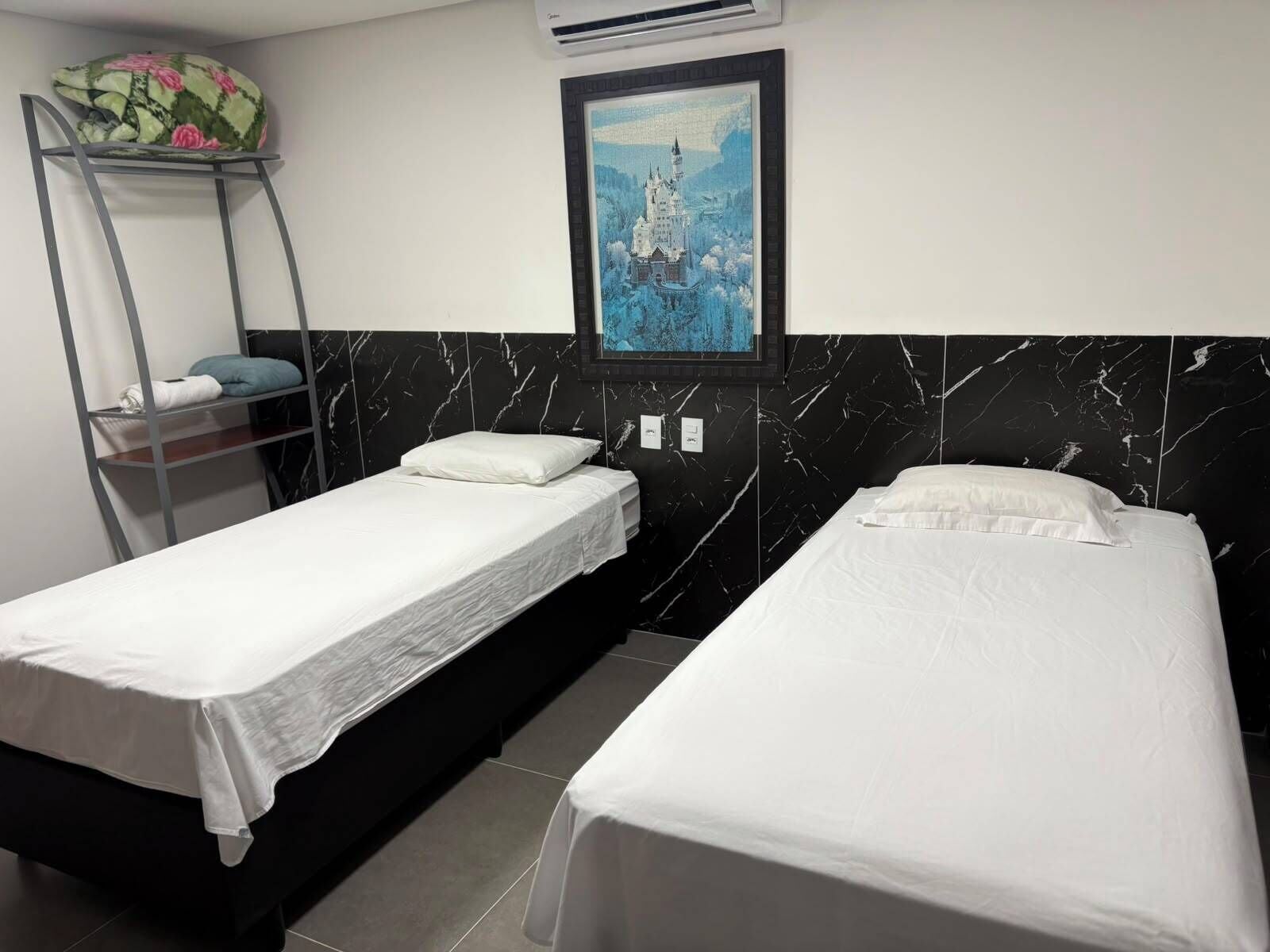 Habitación de hotel con dos camas individuales, pared con diseño de mármol, una pintura de un castillo y un estante metálico con almohadas.