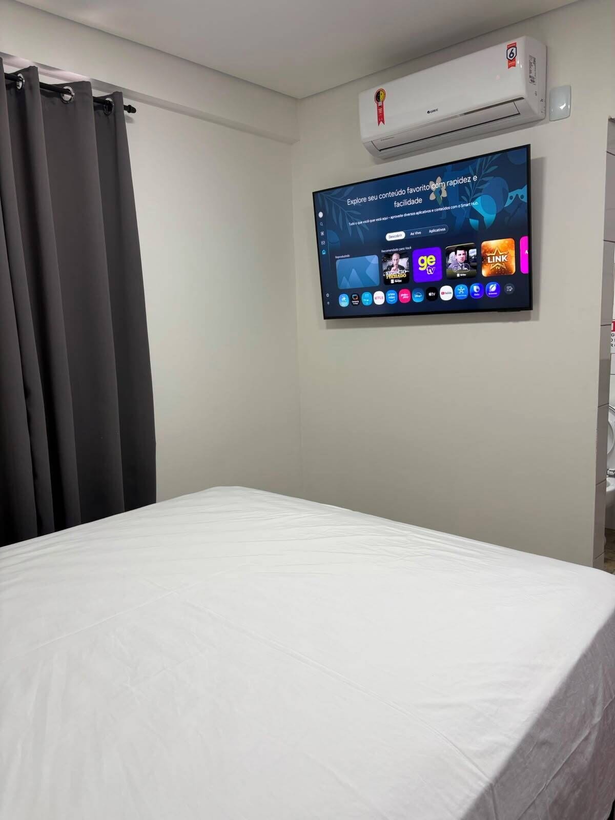 A hotel room with a bed, a flat-screen TV mounted on the wall, and an air conditioning unit above it.