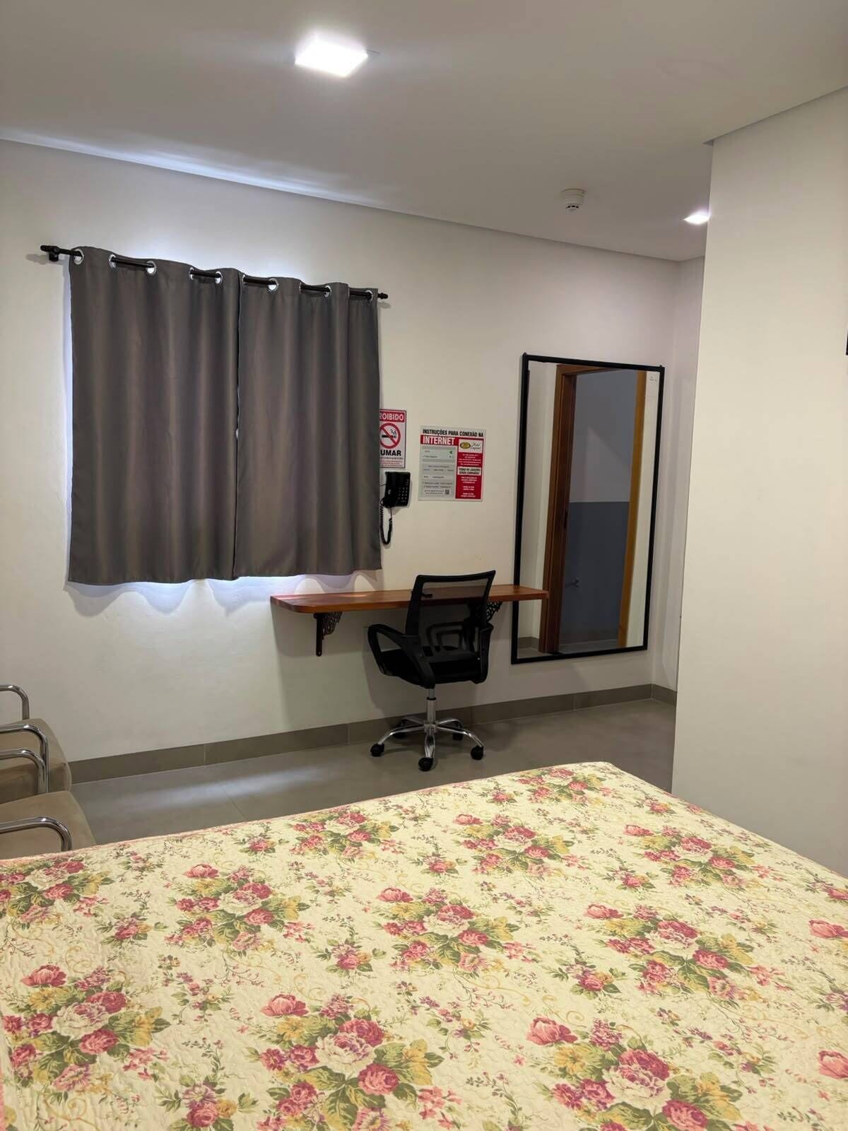 Quarto de hotel com cama, escrivaninha, cadeira, telefone e janela com cortinas.