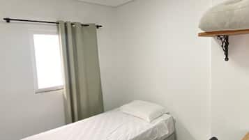 Quarto pequeno, com cama de solteiro, mesa de madeira, luminária no teto e janela com cortinas.