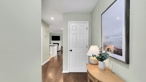 Ein Flur mit Holzboden, einer weißen Tür, einem gerahmten Bild an der Wand und einem Tisch mit Lampe und Pflanze.