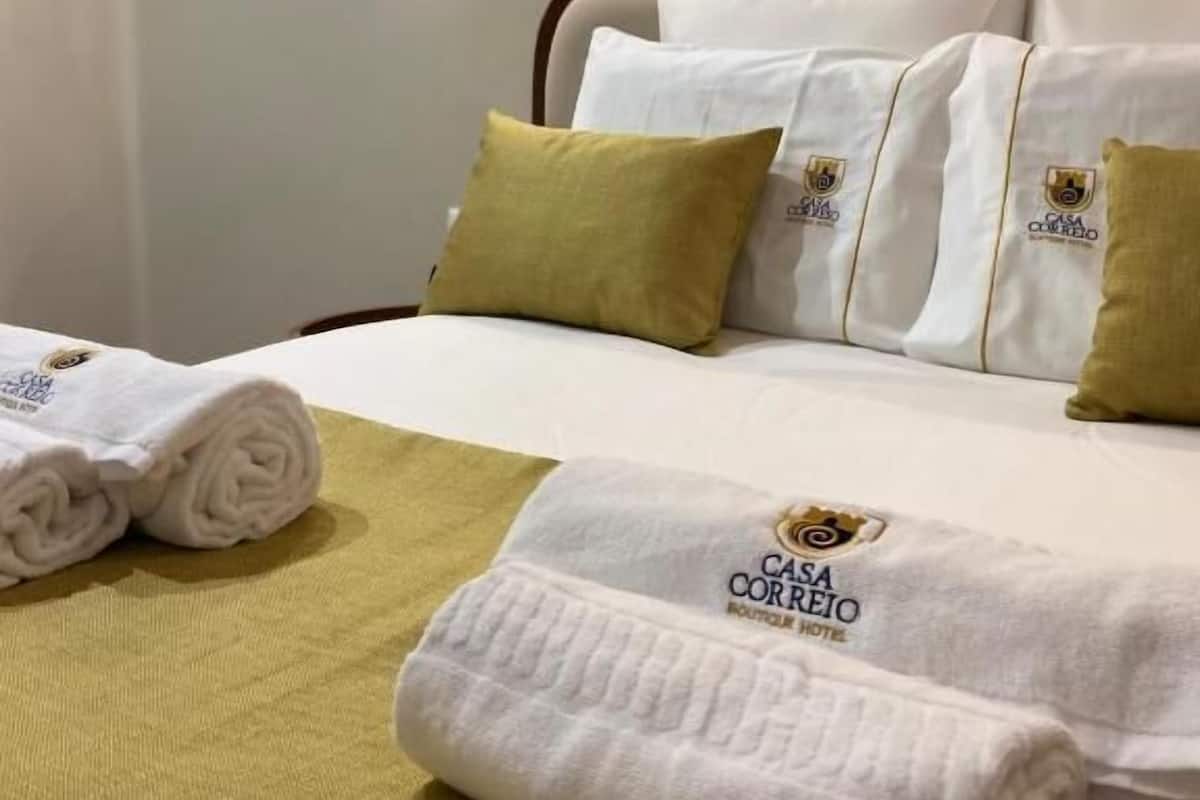 A hotel room with a neatly made bed, white and green pillows, and a rolled towel on the bed.