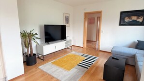 A modern living room with a flat-screen TV, a grey sofa, a yellow rug, and a black ottoman. - Large, cozy vacation apartment in the heart of the Kraichgau. (Kraichtal)