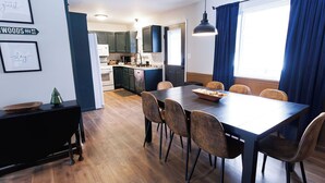 Une cuisine moderne avec des meubles de couleur sombre, un coin repas avec une table et des chaises en bois, et une grande fenêtre ornée de rideaux bleus.
