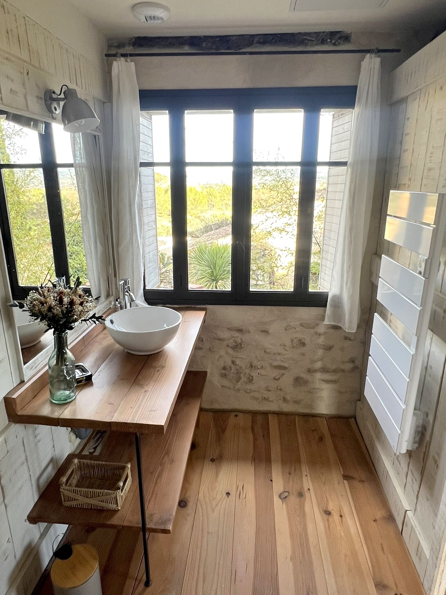 A bathroom with a wooden vanity, a white sink, and a large window with white curtains.