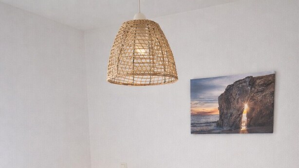 Cama bem arrumada com lençóis e toalhas brancas, luminária pendente de fibra natural e um quadro com pôr do sol sobre um penhasco na parede.
