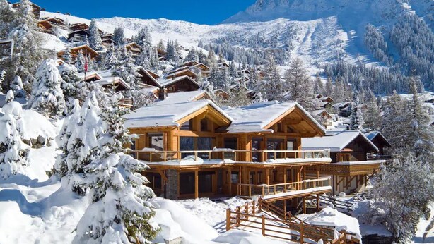Una cabaña nevada con varias cabañas de madera, con montañas cubiertas de nieve al fondo.