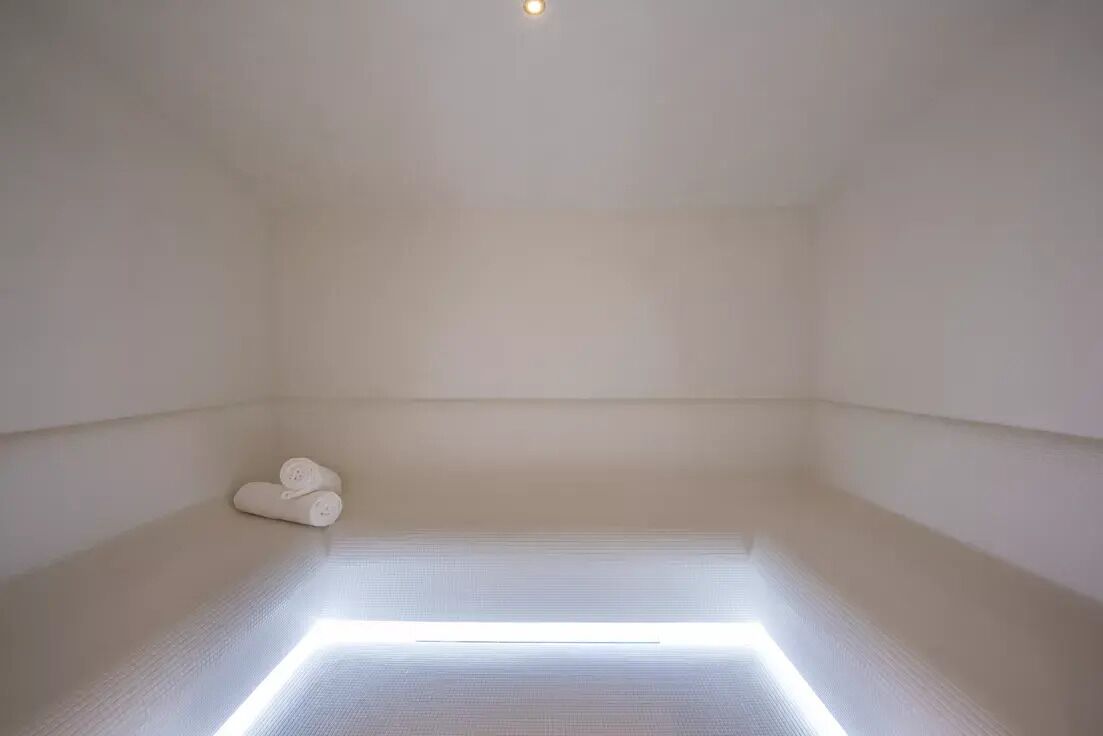 A minimalist room with a geometric light installation on the floor, a small white object on the left, and a single light source on the ceiling.