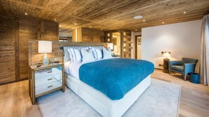 Un dormitorio moderno con techo de madera, una cama grande con manta azul, una mesita de noche con lámpara y una pequeña zona de estar con una silla y una mesita auxiliar.