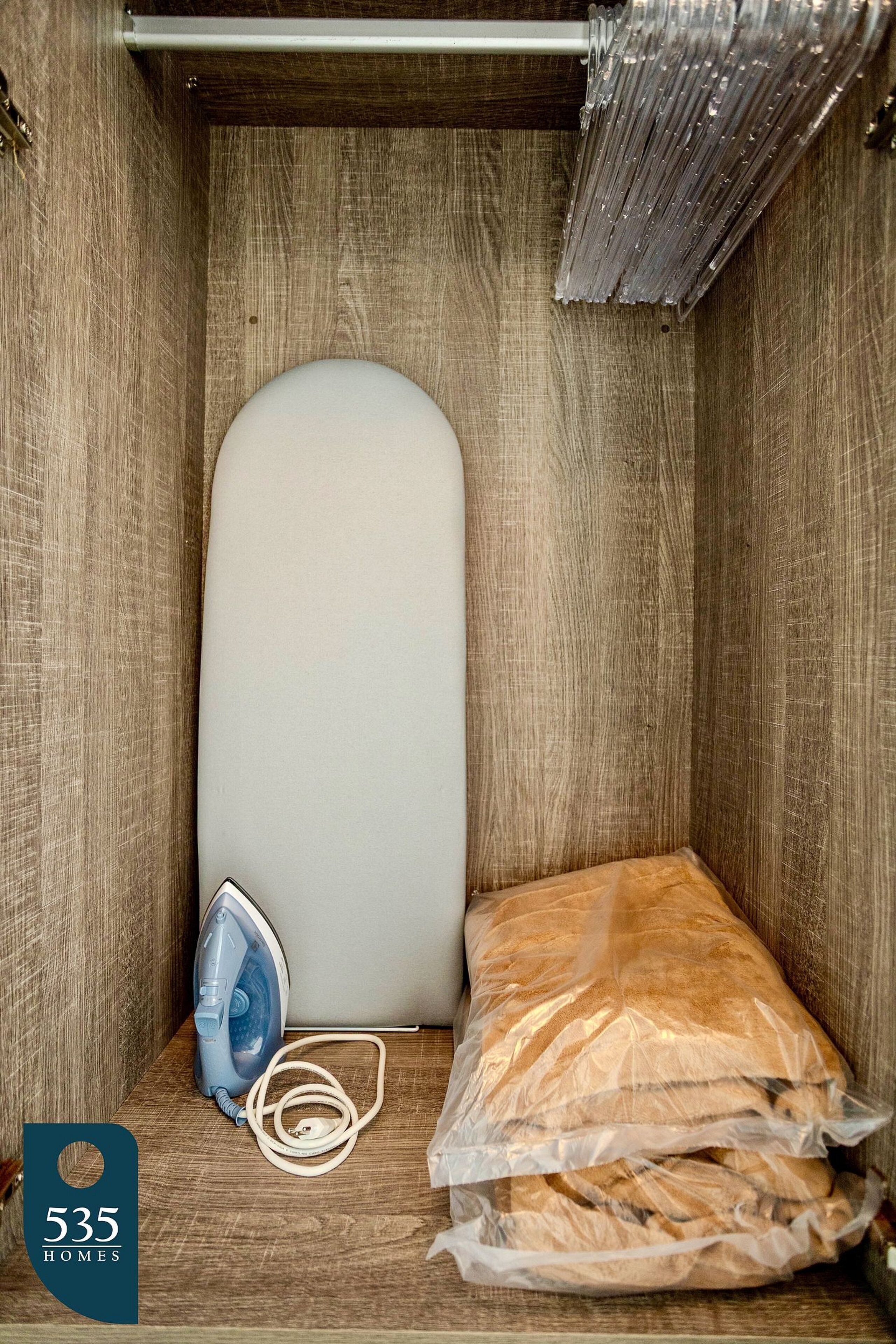 A small, wooden closet with a white ironing board, an iron, and plastic-wrapped bread.