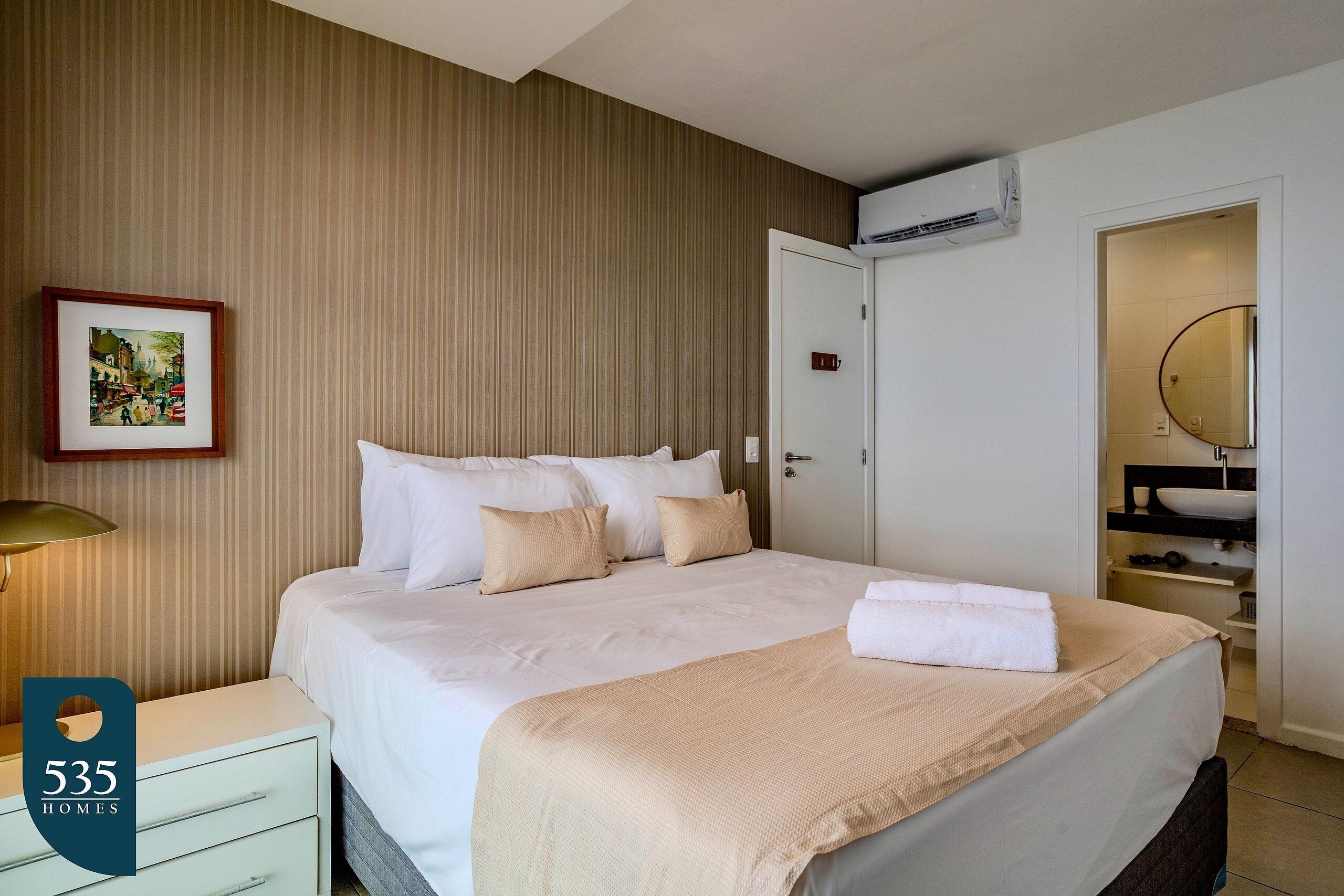 A hotel room with a large bed, a bedside table, a framed picture on the wall, and a bathroom with a sink and mirror.