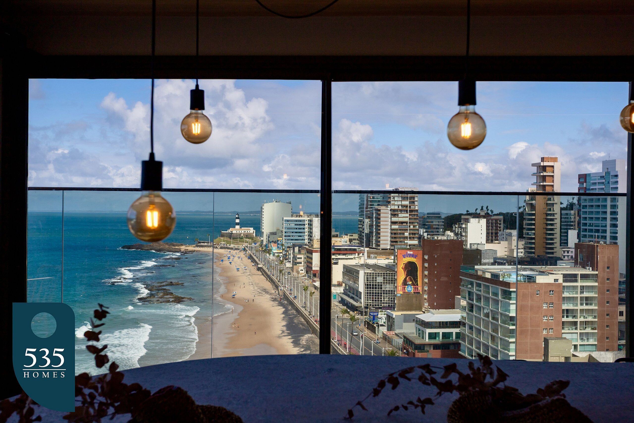 A view from a high-rise hotel room overlooking a beach and cityscape, with hanging light bulbs and a visible sign indicating 535 homes.