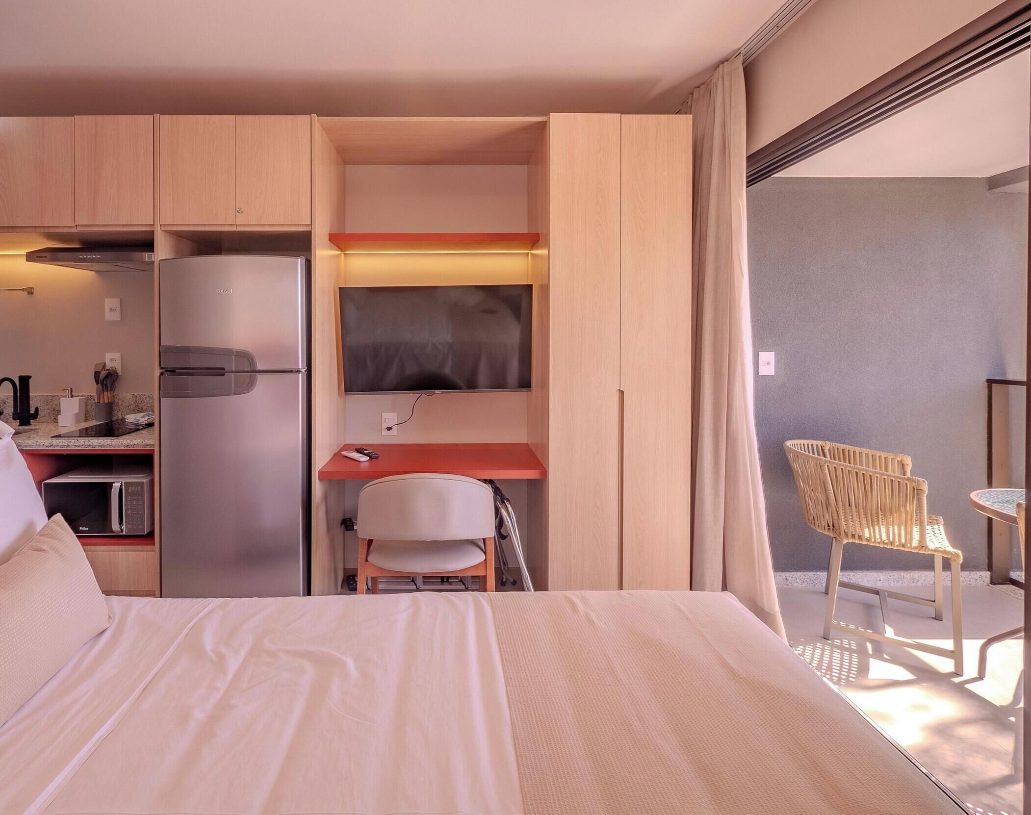 Habitación de hotel con cama, kitchenette, escritorio con silla, televisor y silla de mimbre junto a la ventana.