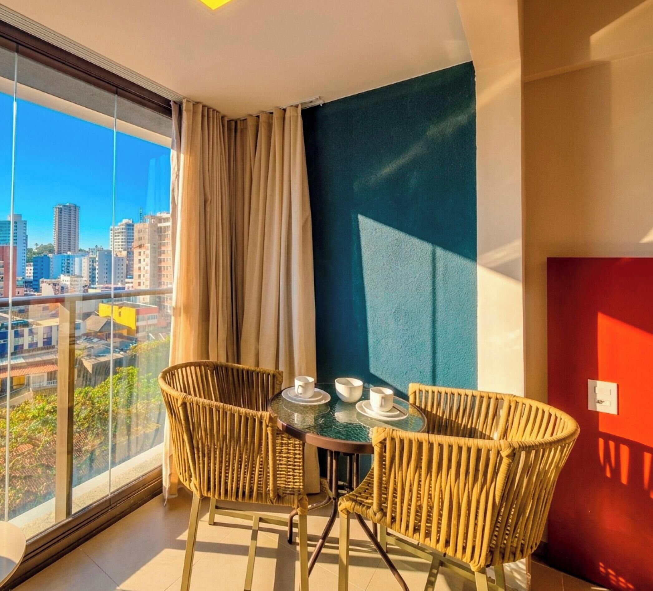 Un balcón con vista a la ciudad, sillas de mimbre, una pequeña mesa con tazas y una pared azul.