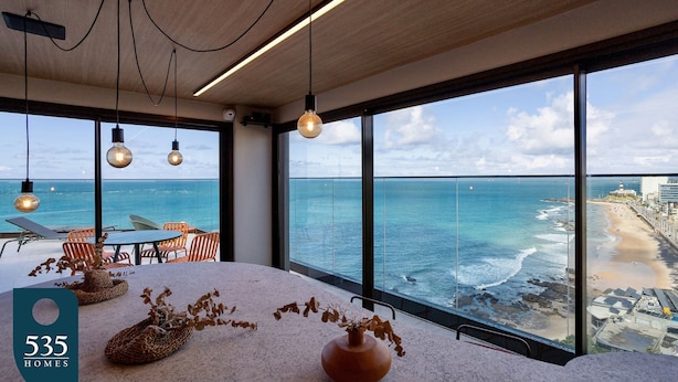 A modern interior with large windows overlooking a beach, a table with chairs, and a vase with dried plants. - Experience Barra’s waterfront with a wide sea view (Salvador)