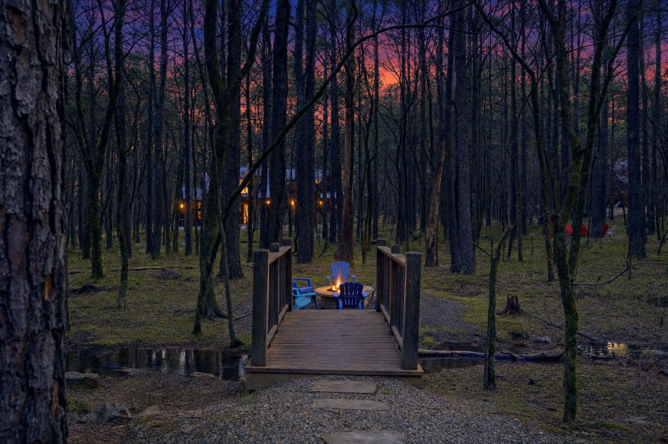 Una terraza de madera que conduce a un espacio de descanso con sillas y una chimenea, rodeada de un área boscosa.
