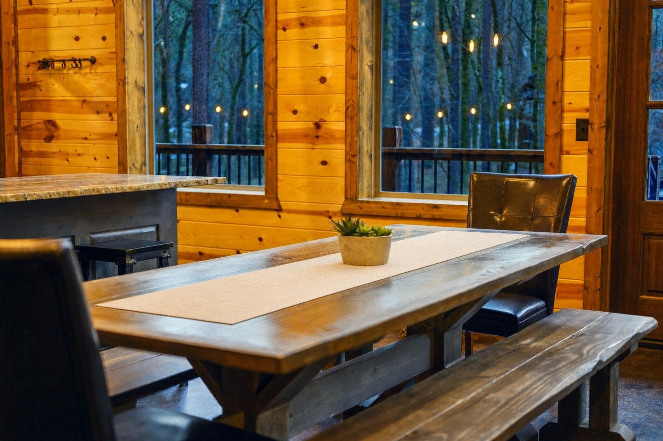 Un interior de madera con una mesa larga y bancos, ventanas grandes con luces colgantes y vistas a árboles.