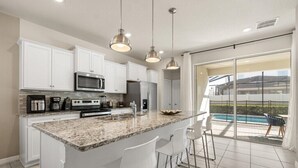 Een moderne keuken met witte kasten, granieten aanrechtbladen en roestvrijstalen apparatuur. Er is een groot eiland met barkrukken en een uitzicht op een zwembad door glazen deuren.