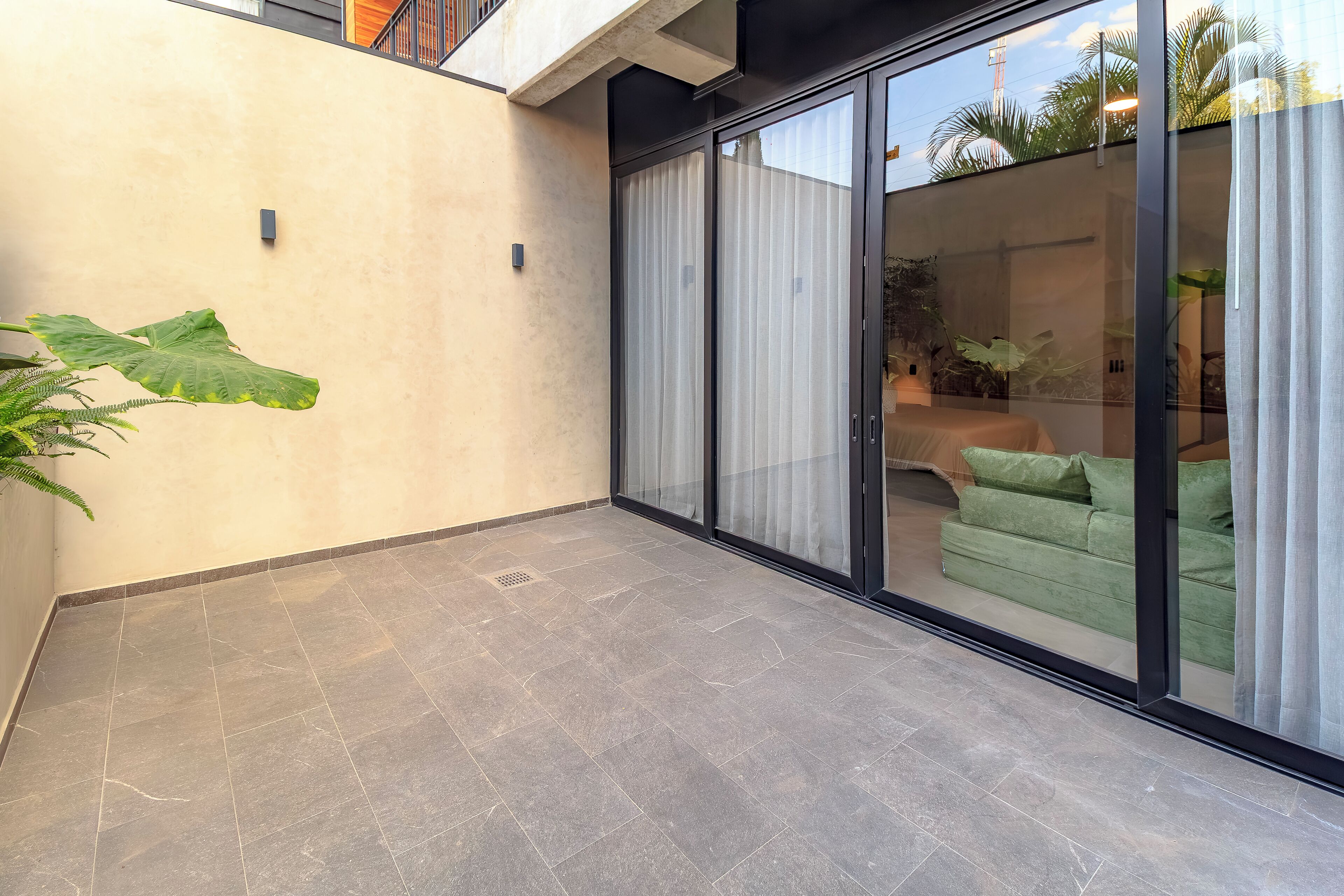 Eine moderne Terrasse mit Fliesenboden, einer großen Grünpflanze und einer Glastür, die zu einem Innenraum mit einem grünen Sofa führt.