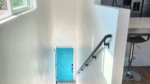 Una scala conduce a una stanza luminosa con una porta blu, una zona cucina con frigorifero in acciaio inossidabile e uno sgabello da bar.