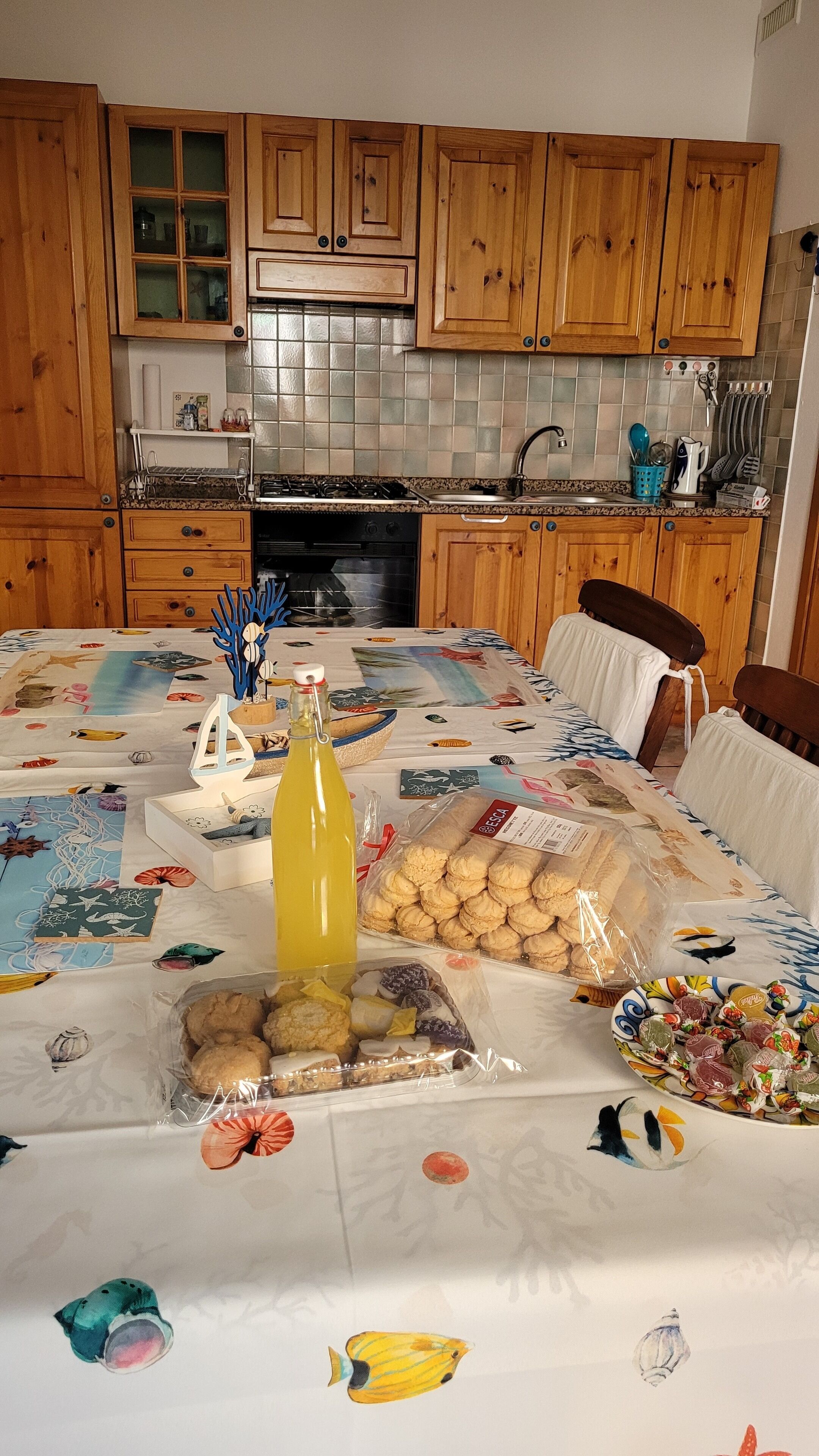 Une cuisine avec des meubles de rangement en bois, un dosseret carrelé et une table à manger recouverte d’une nappe ornée d’illustrations de poissons.