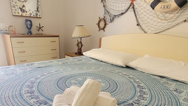 A neatly made bed with a blue patterned bedspread, a stack of white towels, and a life preserver hanging on the netting above the bed. - Villa with garden in Villasimius (Cagliari)