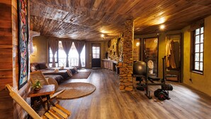 Een interieur van een blokhut met een stenen pilaar, houten vloeren, een bank, een tafel en een fitnessruimte.