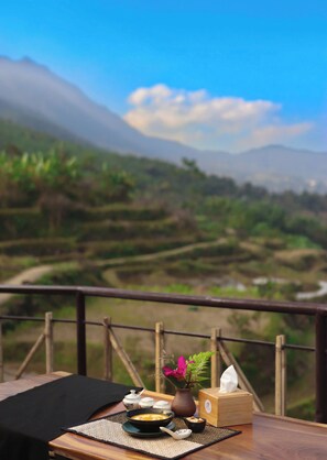 A table with a bowl of soup, a vase with flowers, and a tissue box, set against a scenic mountain view. - Puluong Qui E Ora (Ba Thuoc)