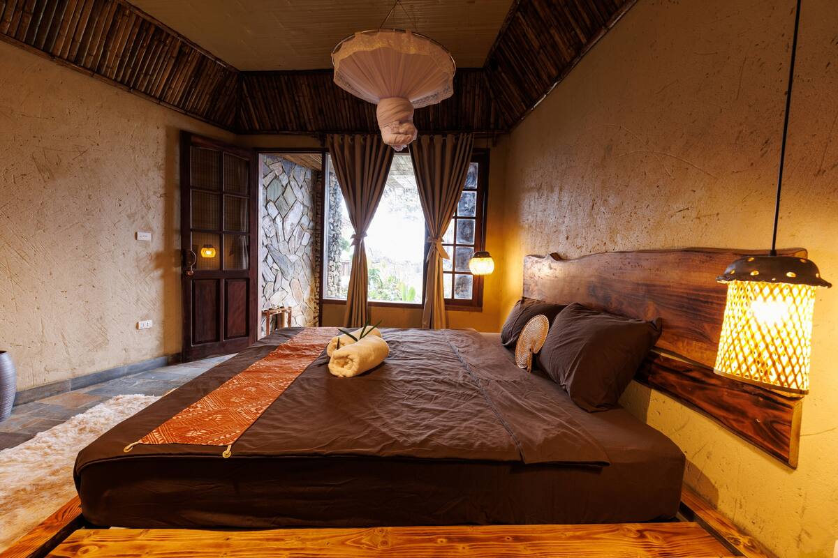 Une chambre à coucher avec un grand lit, une tête de lit en bois, un luminaire suspendu et un mur en pierre.