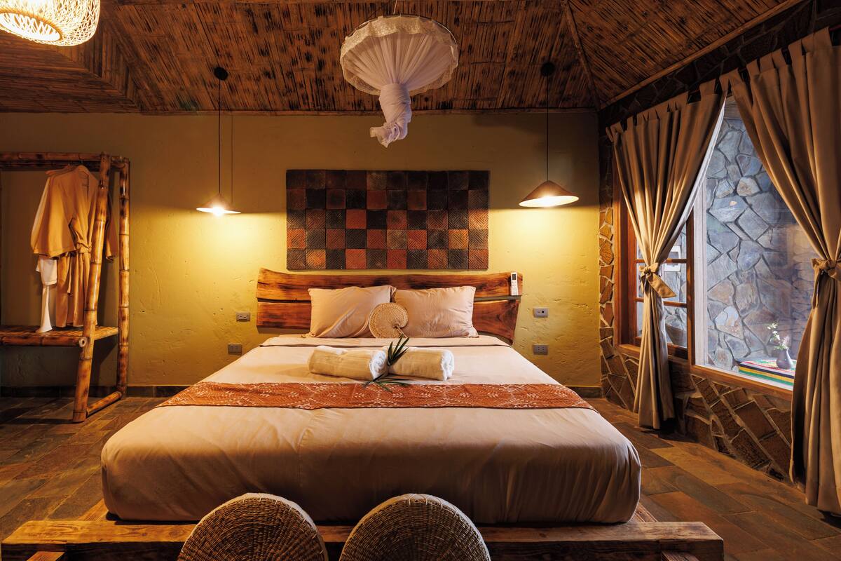 Une chambre avec un grand lit, une tête de lit ornée d’une courtepointe, un luminaire suspendu, un mur en pierre et une fenêtre avec des rideaux.