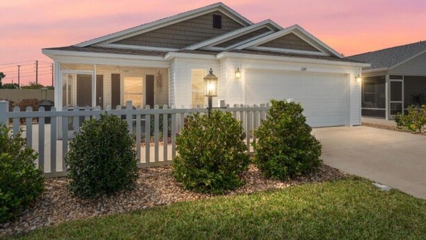 A single-story house with a white exterior, a covered porch, and a fenced yard with small bushes. - Centrally Located close to Brownwood, Sawgrass and Eastport with Golf Cart. (The Villages)