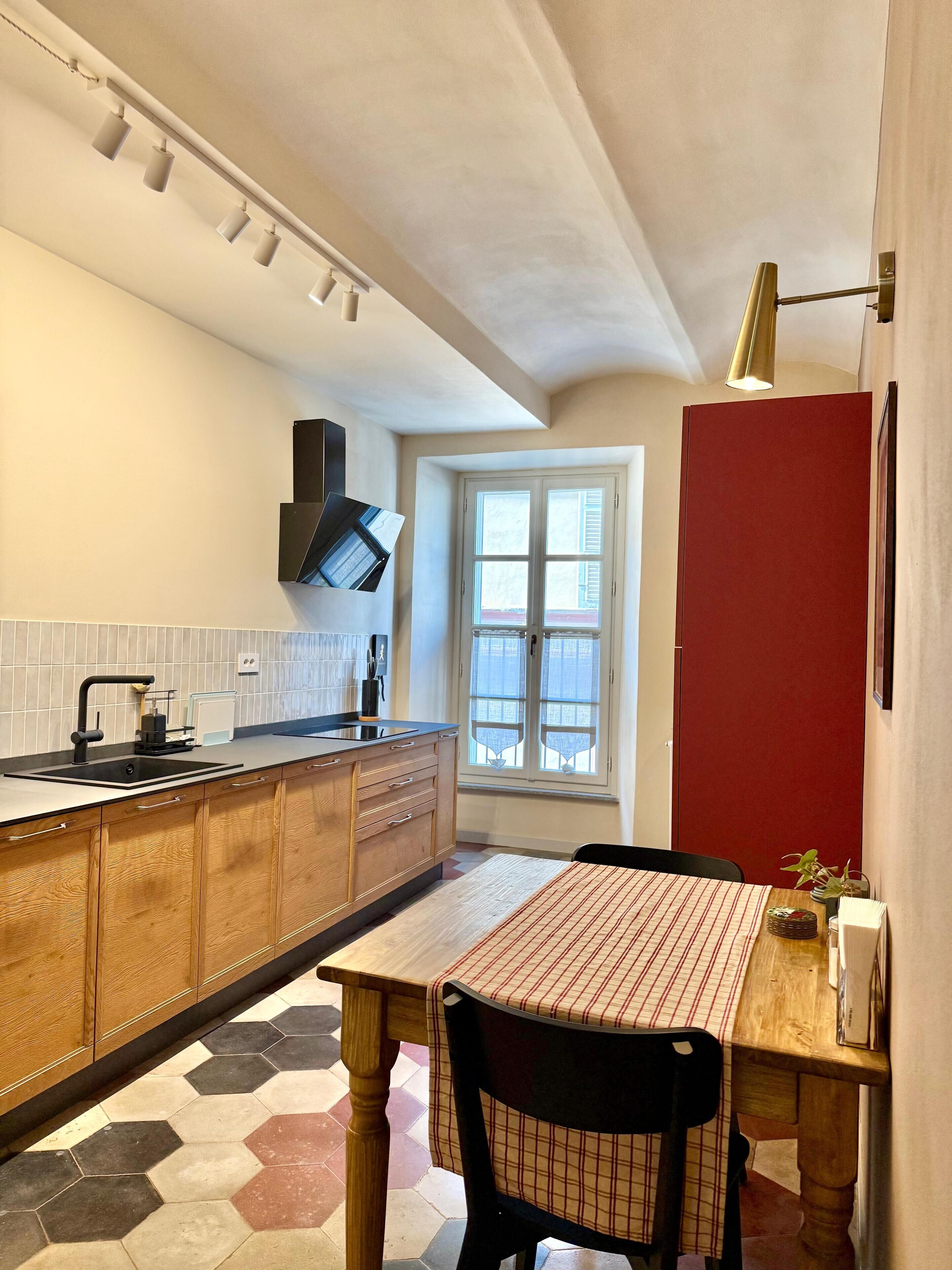Une cuisine avec des armoires en bois, un plan de travail noir, une fenêtre et une porte rouge.