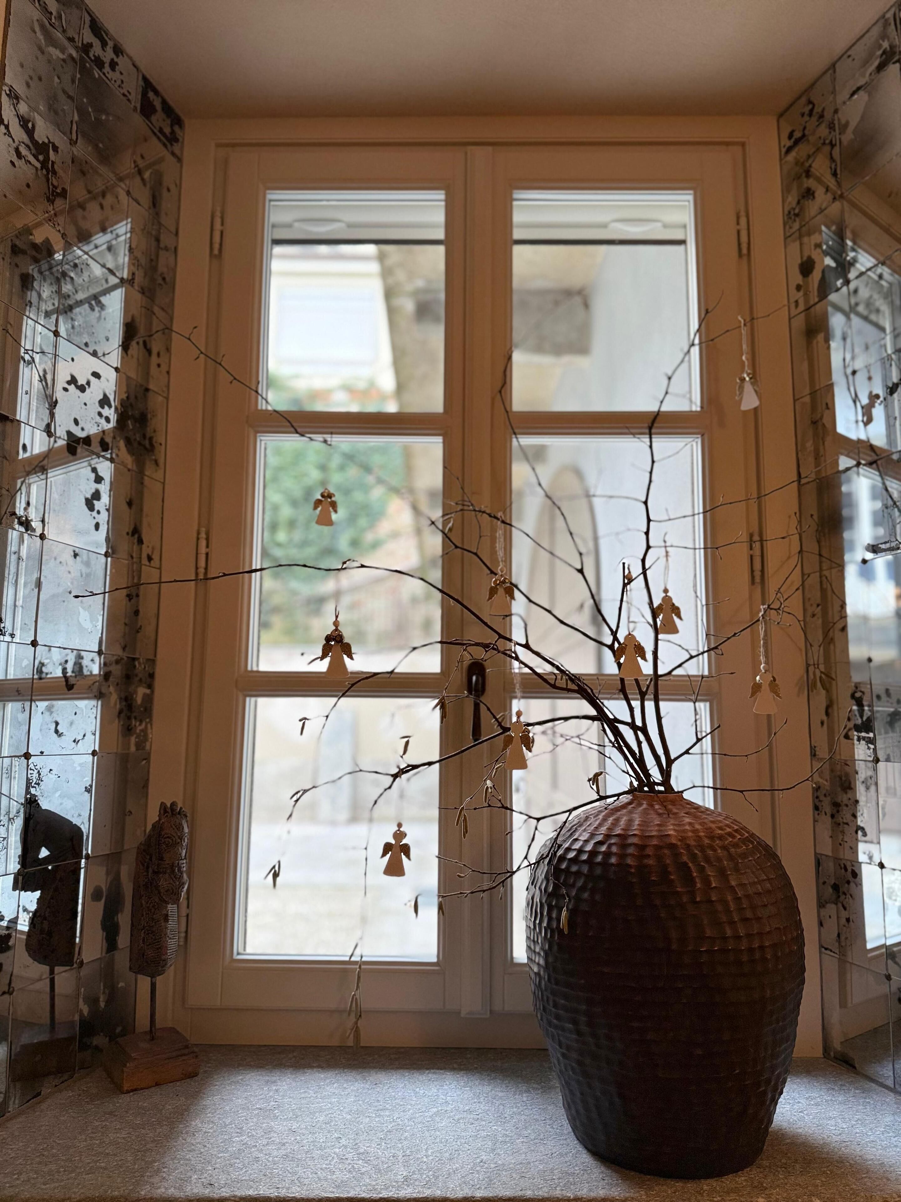 Ein Zimmer mit großem Fenster, einer strukturierten Vase mit Zweigen und einer kleinen Statue auf der Fensterbank.