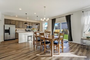 A modern kitchen with a central island, wooden dining table with chairs, pendant lighting, and a view of the backyard through large windows. - New Spacious and Luxurious Home (Colorado Springs)