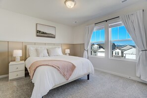 A bedroom with a large bed, two bedside tables with lamps, a headboard, a framed picture on the wall, and a view of houses through the window. - New Spacious and Luxurious Home (Colorado Springs)