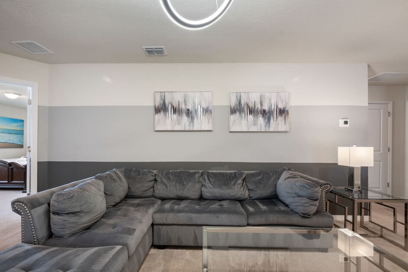 Een moderne woonkamer met een grijze modulaire bank, twee abstracte schilderijen, een glazen salontafel en een lamp op een bijzettafeltje.