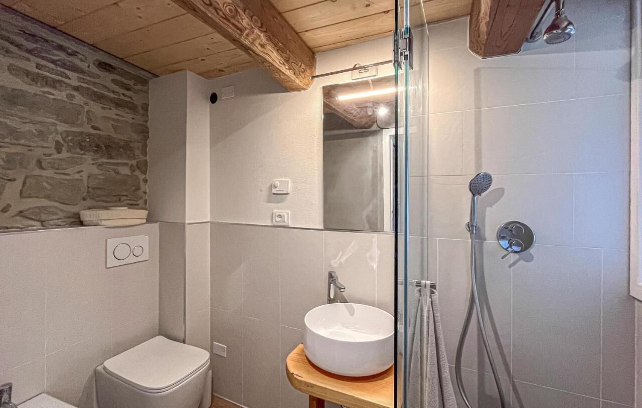 A bathroom with a stone wall, a wooden ceiling, a white sink, a glass shower enclosure, and a toilet.