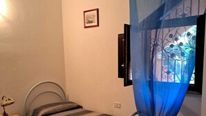 Een kleine slaapkamer met een bed, een bureau en een stoel. Er is een raam met een blauw gordijn en een foto aan de muur.