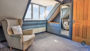 Une chambre avec un plafond en pente, une grande fenêtre, une chaise, une commode et une porte.