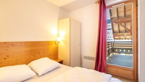 A hotel room with a wooden headboard, a bed with white bedding, a nightstand, a wall-mounted light, and a door leading to an outdoor wooden deck. - Pet-friendly Family Home With Balcony (Saint-Sorlin-d'Arves)
